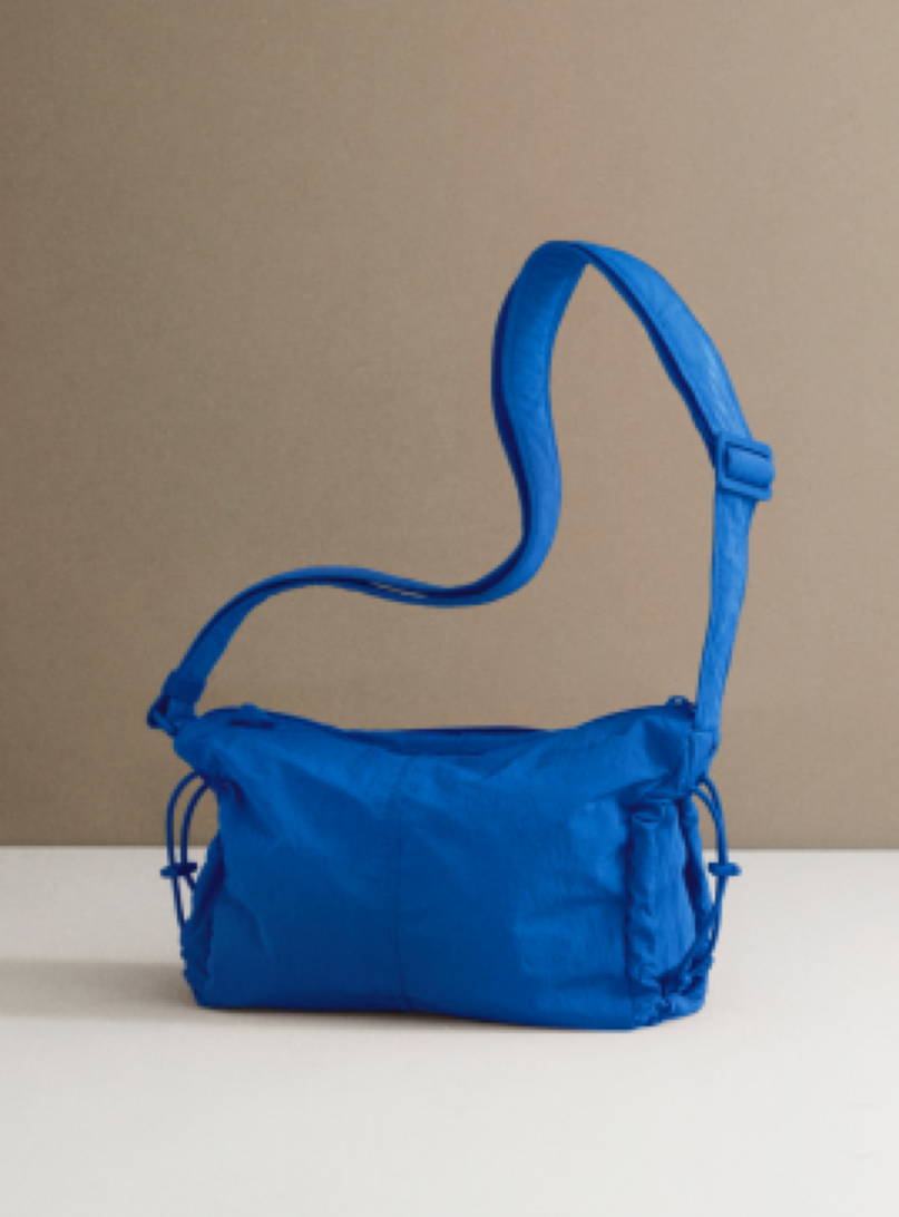 A bright blue nylon shoulder bag with a wide adjustable strap and drawstring details, displayed on a white surface against a neutral beige background.