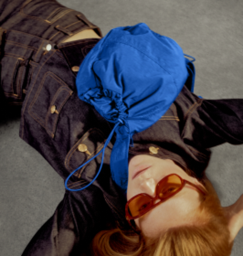 A person lying on a gray floor wearing a bright blue drawstring pouch bag across their chest, with dark denim clothing and orange-tinted sunglasses visible.