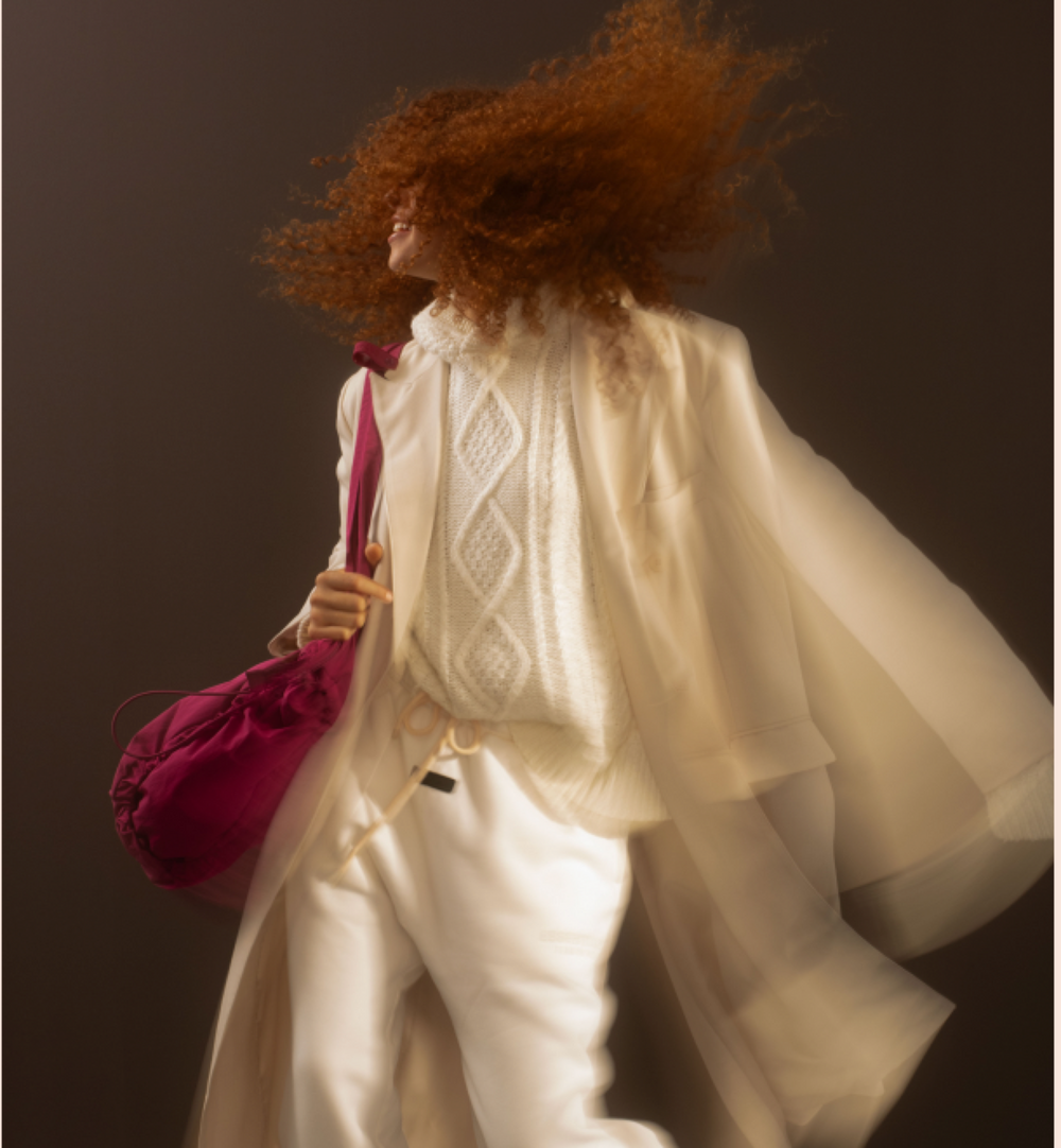 A person with voluminous curly red hair is captured mid-motion wearing an oversized cream-colored sheer cape, a cream knit sweater, and cream trousers, while carrying a bright magenta drawstring bag against a dark brown studio background.