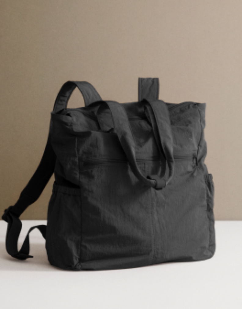 A black fabric tote bag with dual shoulder straps, shown standing upright on a white surface against a warm beige background.