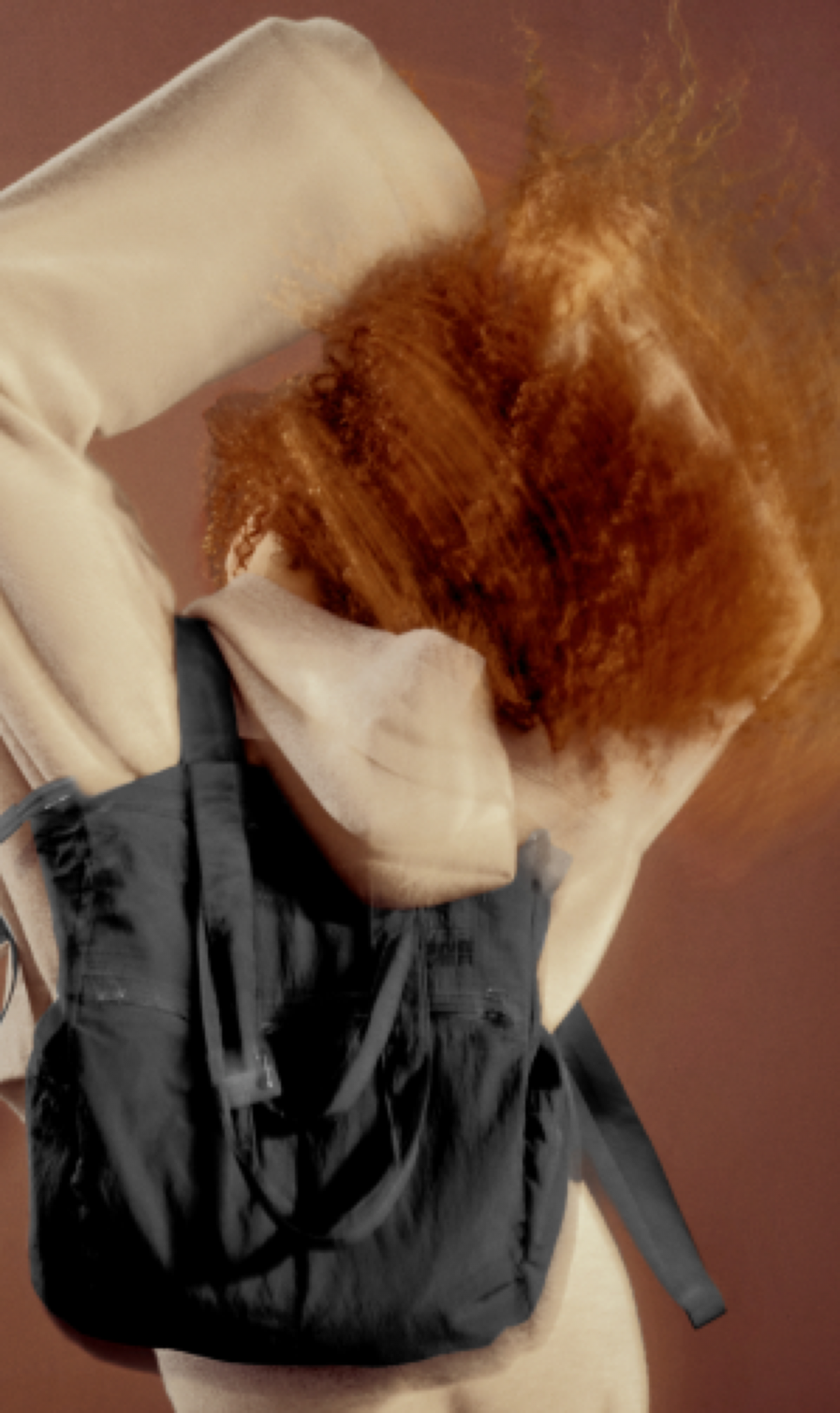 A person with voluminous curly red hair wearing a cream-colored hoodie and carrying a black canvas backpack, set against a solid warm brown background.