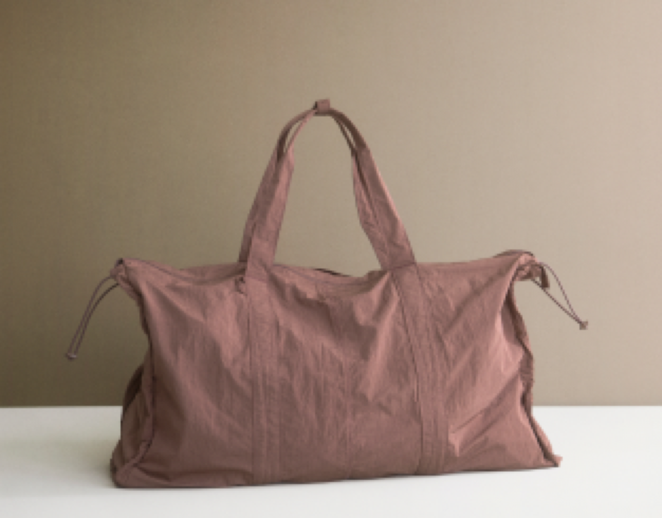 A dusty rose or mauve-colored canvas duffel bag with dual top handles and drawstring ties, resting on a white surface against a solid beige background.