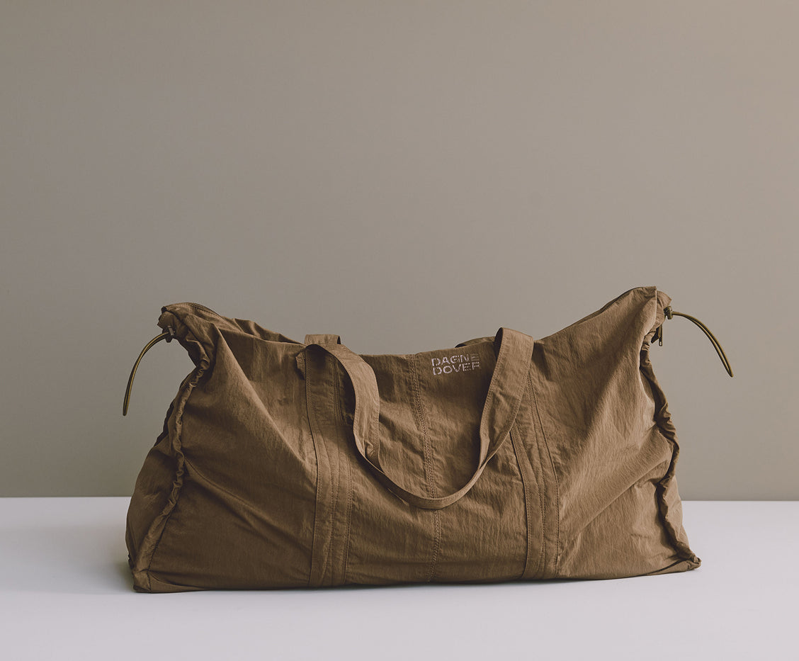 A large olive brown duffel bag with dual shoulder straps and drawstring ties, displayed on a white surface against a neutral gray background. The brand name 'DAGNE DOVER' is printed on the side.