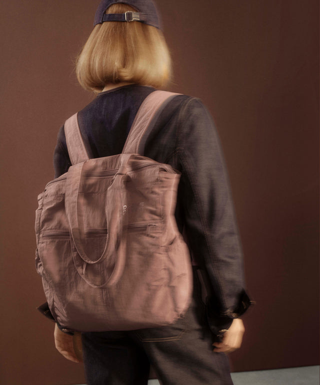 A person viewed from behind wearing a dusty mauve-brown canvas tote bag over their shoulders, styled with a dark cap, dark jacket, and dark pants against a solid dark brown background.