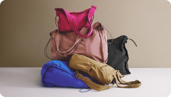 A stack of four different travel and duffel bags in various colors including pink, dusty rose, blue, and tan, arranged on a white surface against a solid beige background.