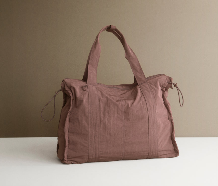 A dusty rose canvas duffel bag with dual top handles and drawstring details, displayed on a white surface against a solid taupe background.