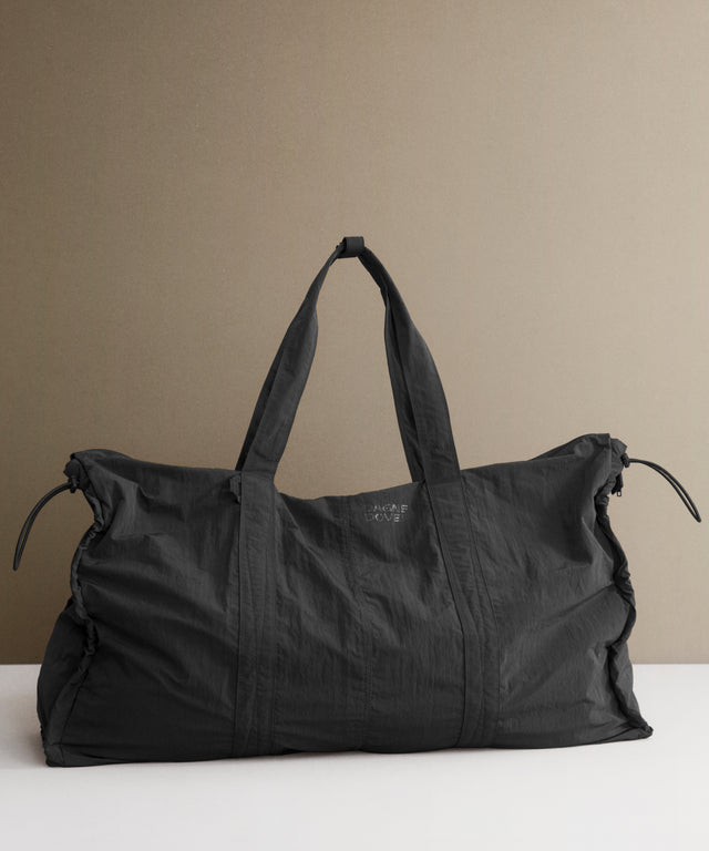 A large black duffel bag with dual shoulder straps, resting on a white surface against a warm beige background.