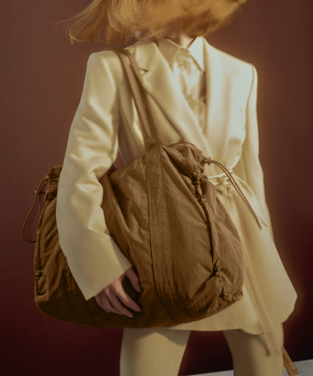 A person in a cream-colored blazer and matching trousers carries a large brown drawstring bag over their shoulder against a dark reddish-brown background.