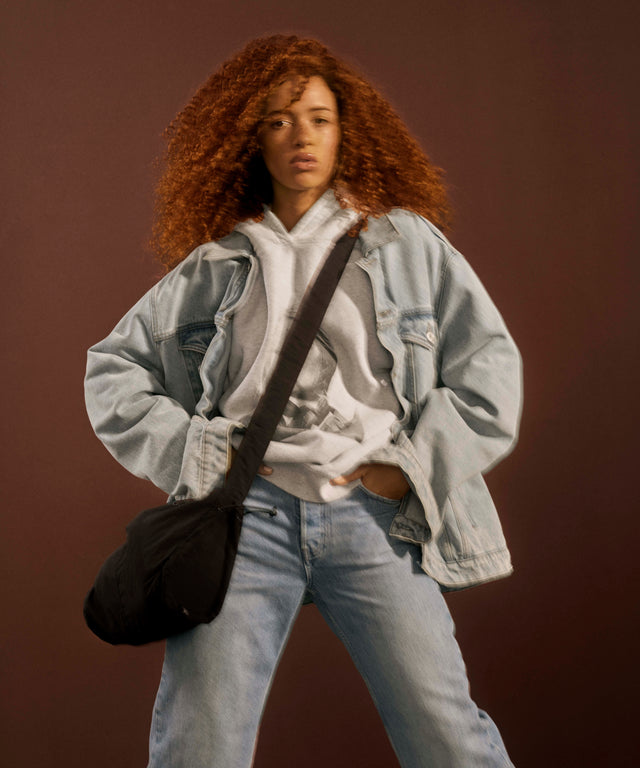 A model with voluminous curly red hair is wearing a light-wash denim jacket over a white hoodie, paired with light blue jeans and a black crossbody bag, set against a solid dark brown studio background.