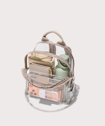 Clear bag with beige straps and a front pocket holding everyday items on a white background. Stadium compliant.