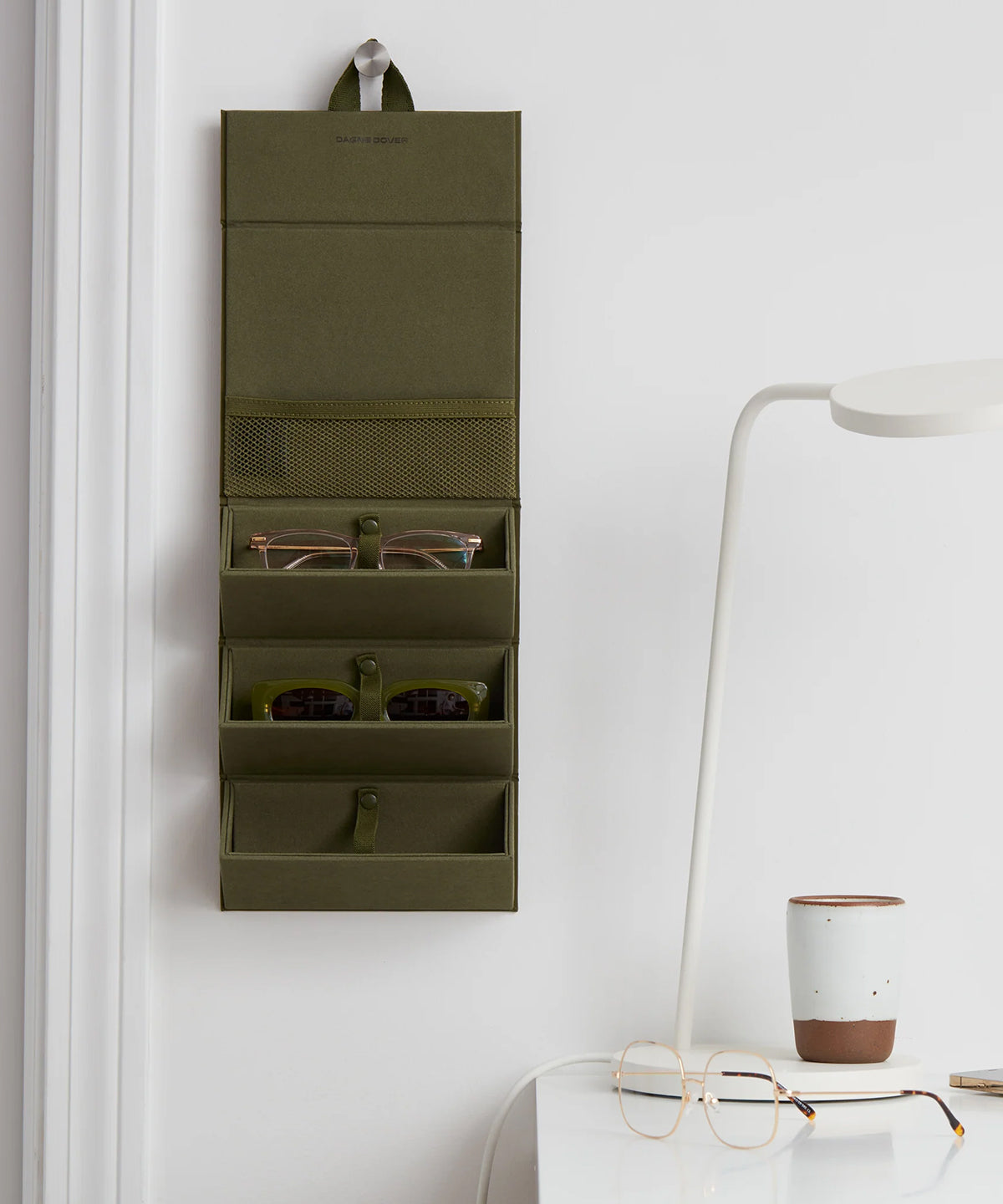 Dagne Dover Remi foldable glasses holder in dark green hung up and holding glasses by a desk.
