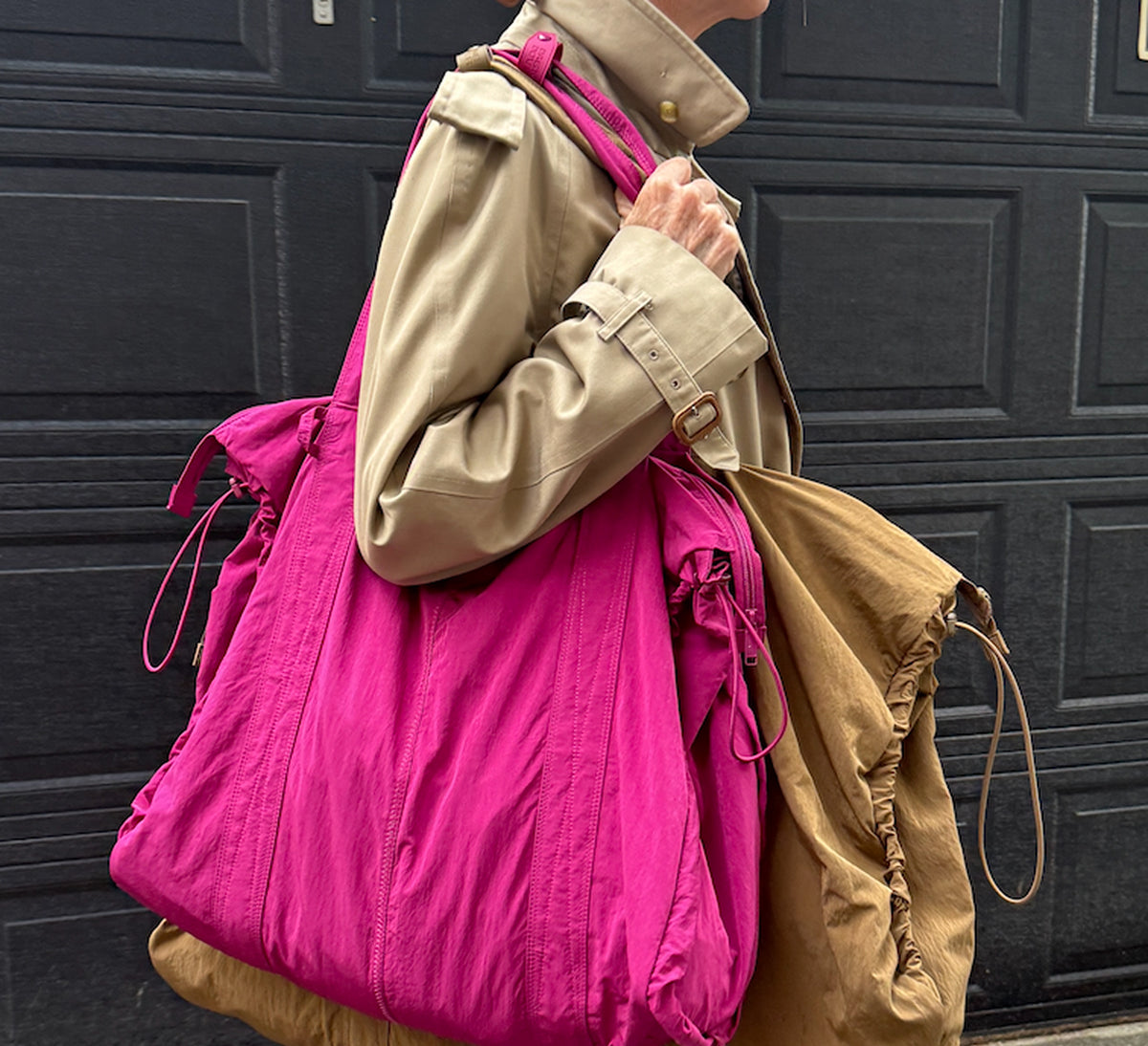 A person wearing a beige trench coat carries a large bright pink tote bag over one shoulder, with a tan drawstring bag slung over the other shoulder, in front of a dark paneled door.