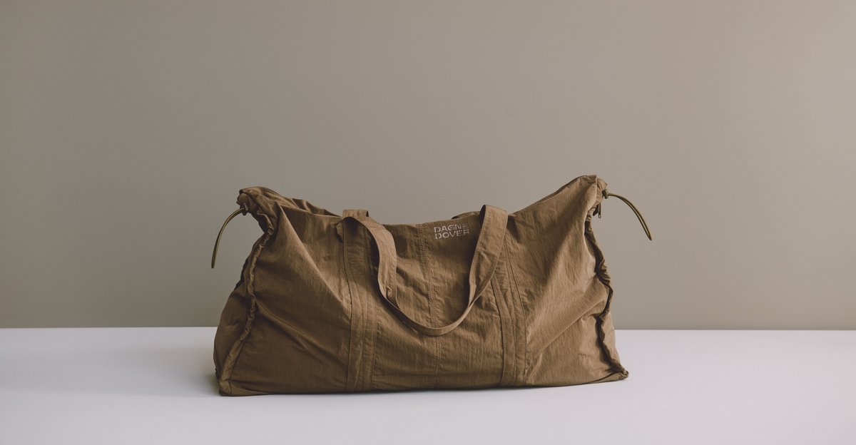 A brown crinkled fabric duffel bag with drawstring handles sits on a white surface against a solid beige background.