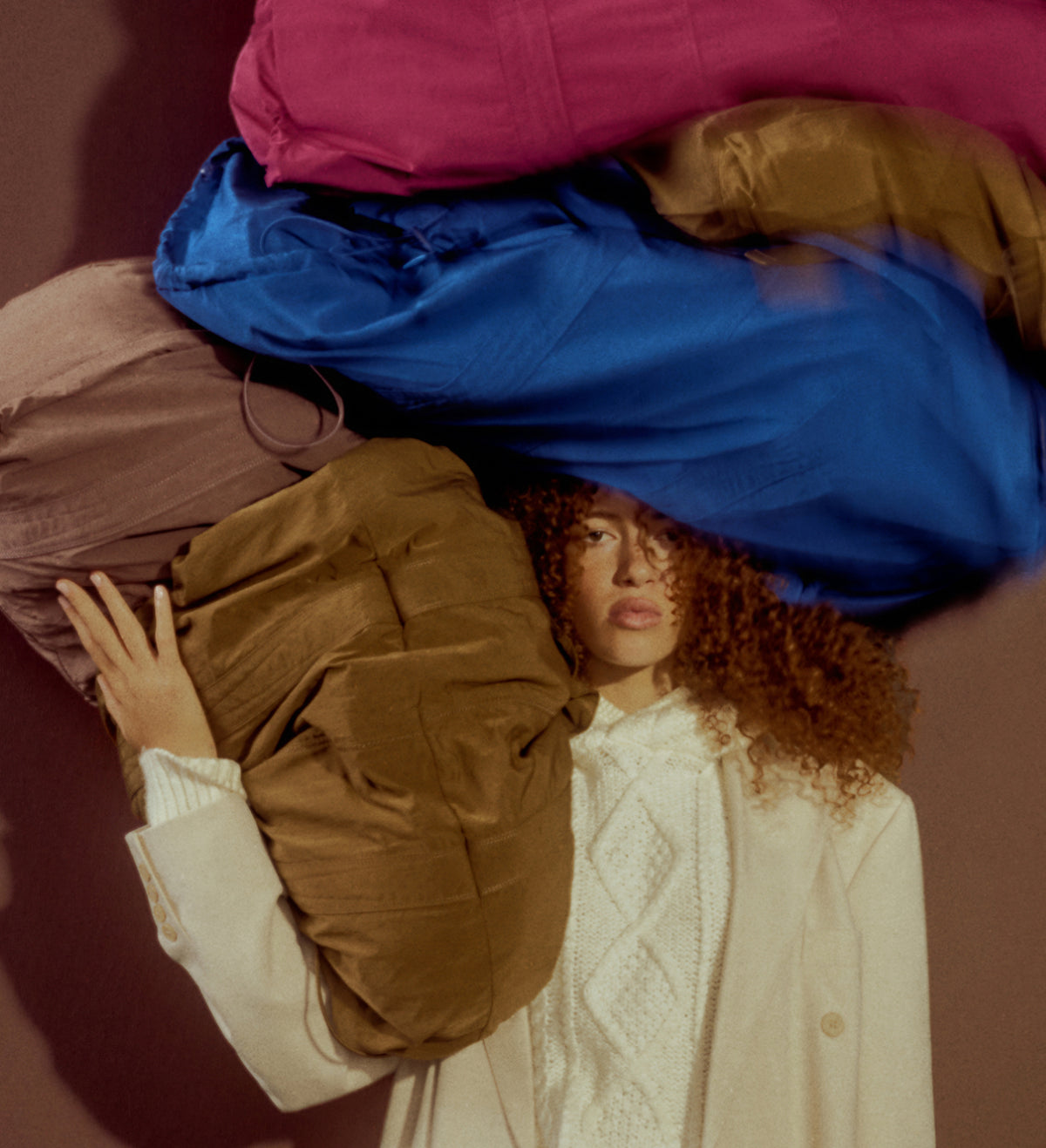 A person with curly hair wearing a cream knit sweater is posed under a large stack of oversized colorful bags in pink, blue, and brown against a dark brown background.