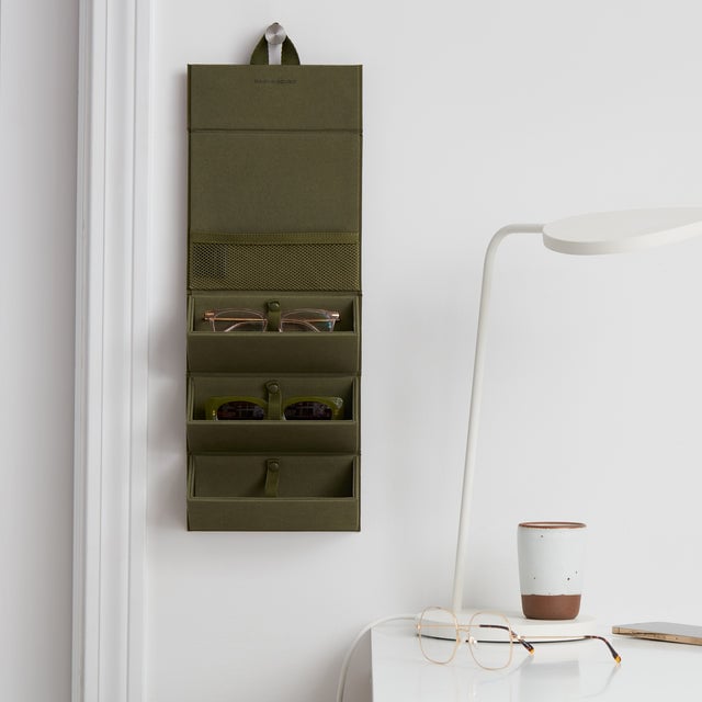 Dagne Dover Remi foldable glasses holder in dark dark green hung up and holding glasses next to a desk.