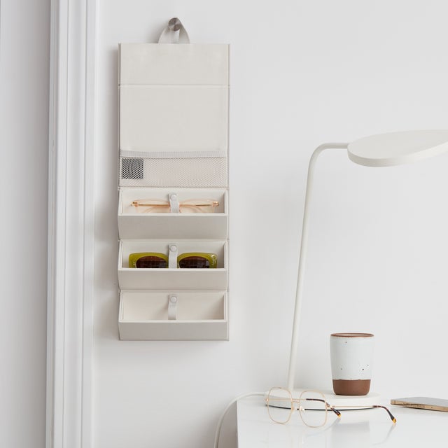 Dagne Dover Remi foldable glasses holder in white hung up and holding glasses next to a desk. 